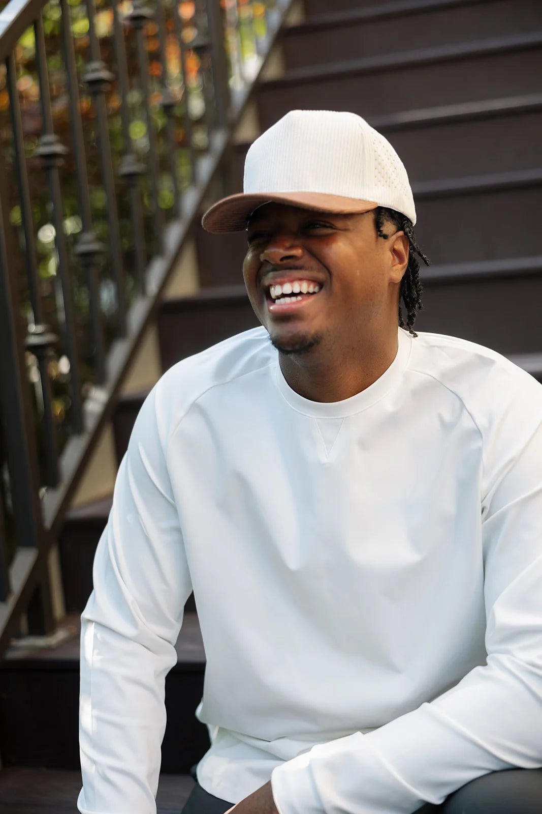 Laughing model wearing a cream crewneck and cap while seated outdoors