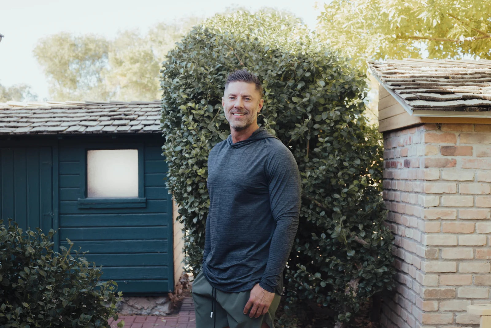 Smiling model wearing a navy hoodie outdoors beside a garden building