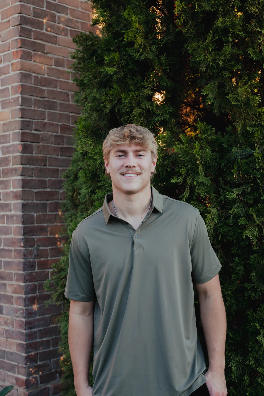 Smiling model wearing an olive polo beside brick and greenery